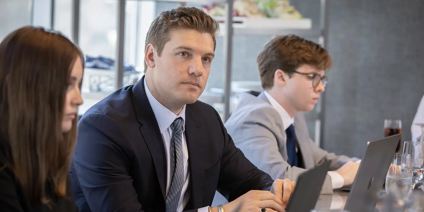 A professional man in a dark suit and tie sits at a conference table with colleagues, looking attentively toward a speaker during a business meeting.