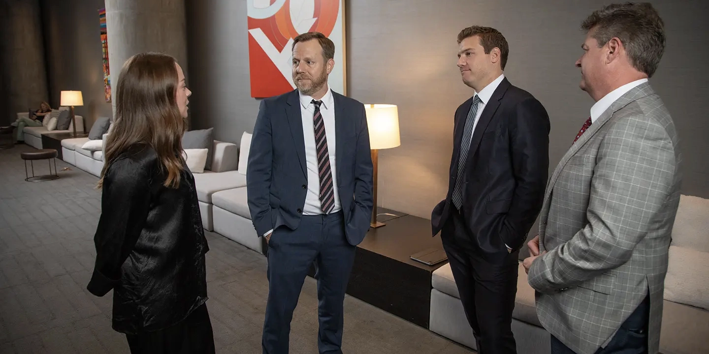 A group of four business professionals, three men and one woman, standing together and talking in a modern lobby.
